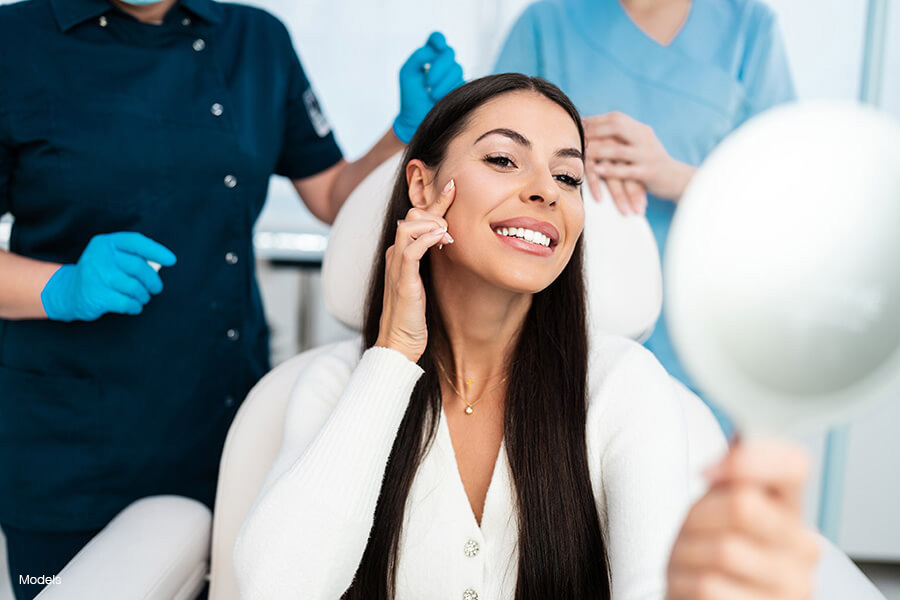 A beautiful smiling woman admires her youthful face in a handheld mirror while two medical professionals stand behind her.